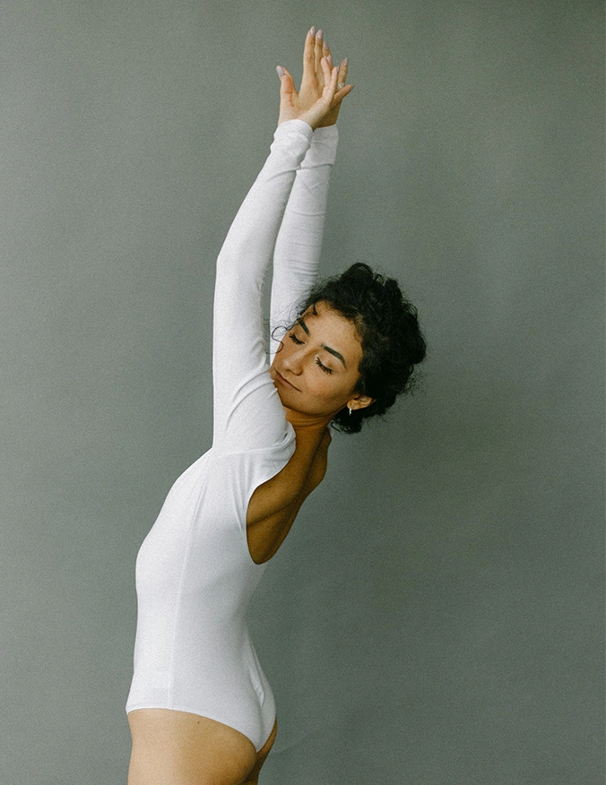 Woman in white bodysuit performing graceful dance stretch with arms raised overhead against grey background - Laceration repair in Parsippany, NJ.