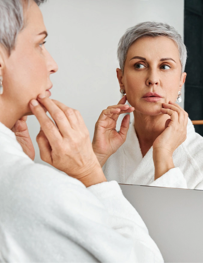 Woman with gray hair examining her face in mirror while touching jawline in white bathrobe - skin cancer reconstruction in Parsippany, NJ.