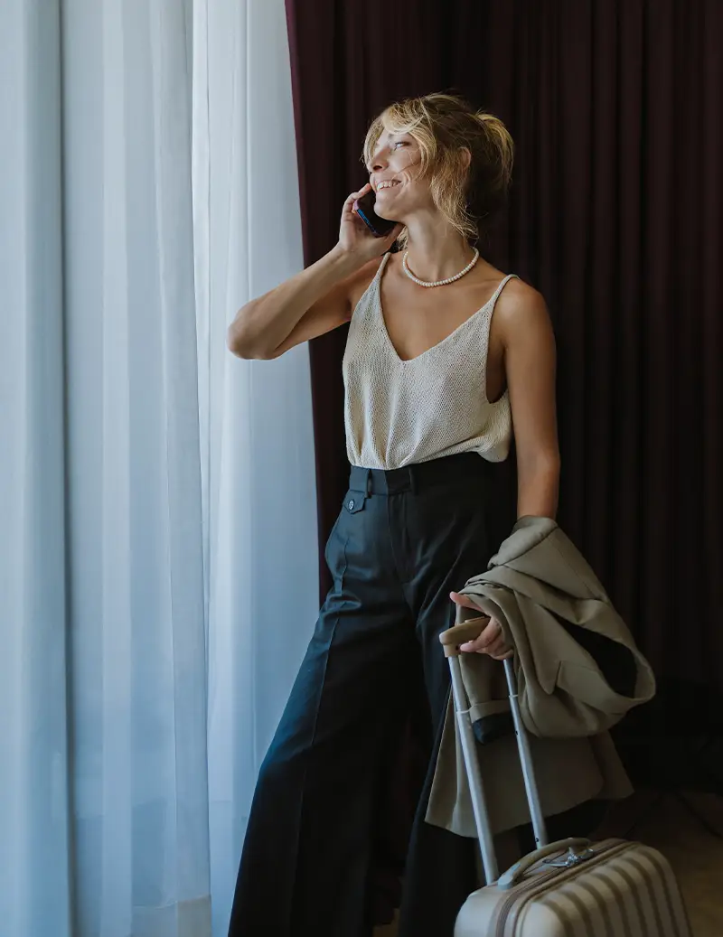 Woman on phone wearing white tank top and dark pants standing by curtained window with luggage