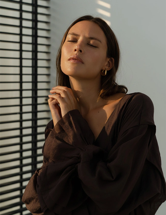 Woman in black blouse posing thoughtfully near window blinds with dramatic natural lighting and shadows - Chin liposuction in Parsippany, NJ.