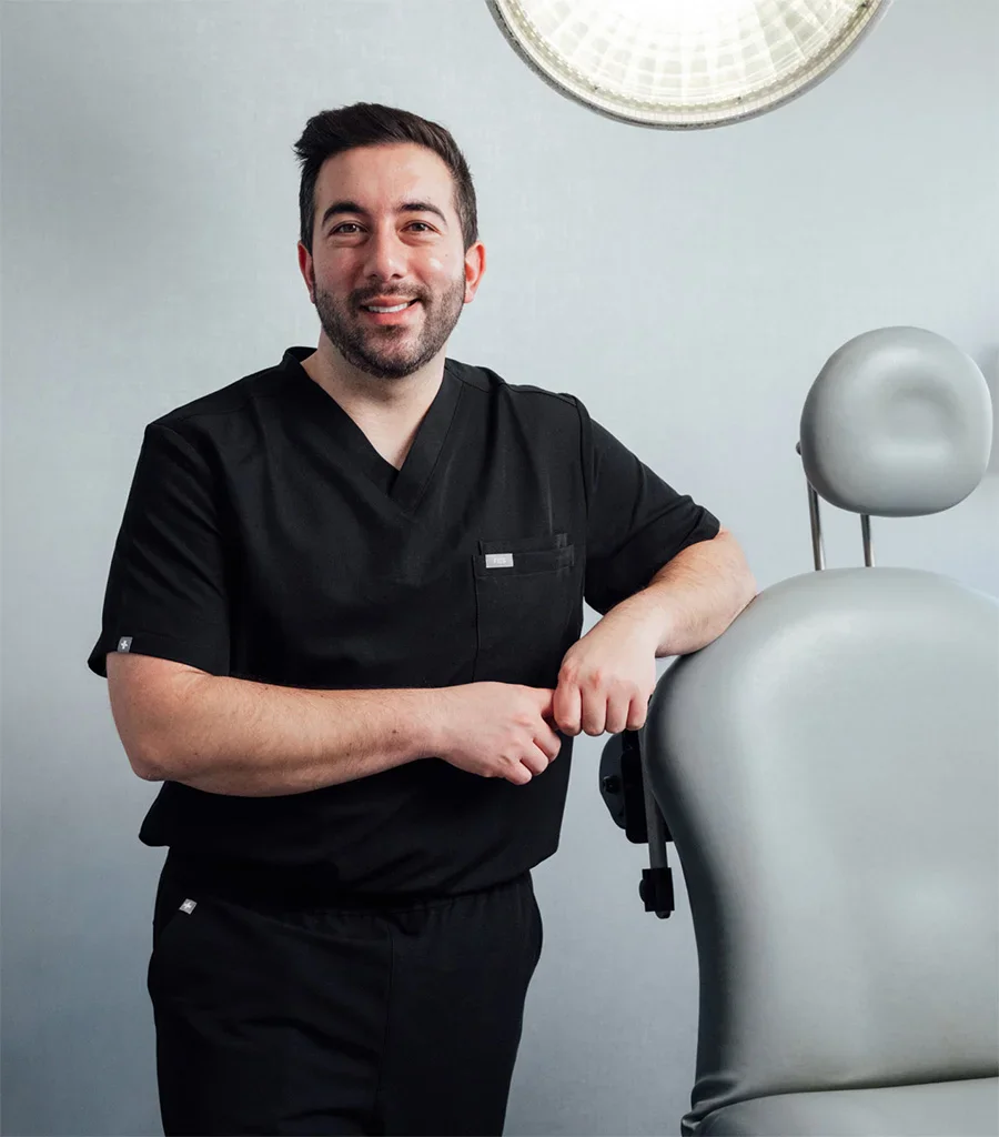 Doctor Shuja Shafqat in black scrubs smiling beside medical training mannequin in clinical office setting