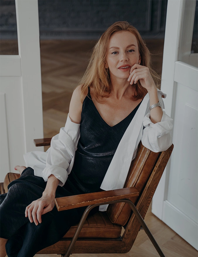 Woman with red hair in black dress and white cardigan sitting in wooden rocking chair on porch - facial fat transfer in Parsippany, NJ.