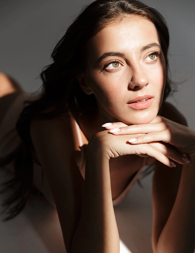 Woman with dark hair resting chin on hands in natural sunlight with soft shadows on face - skincare in Parsippany, NJ.